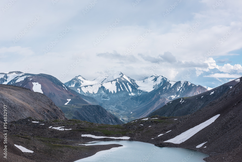 Dramatic view to most beautiful blue alpine lake against snow-covered range with few pointy peaks. Atmospheric landscape with azure mountain lake against three snowy peaked tops under gray cloudy sky.