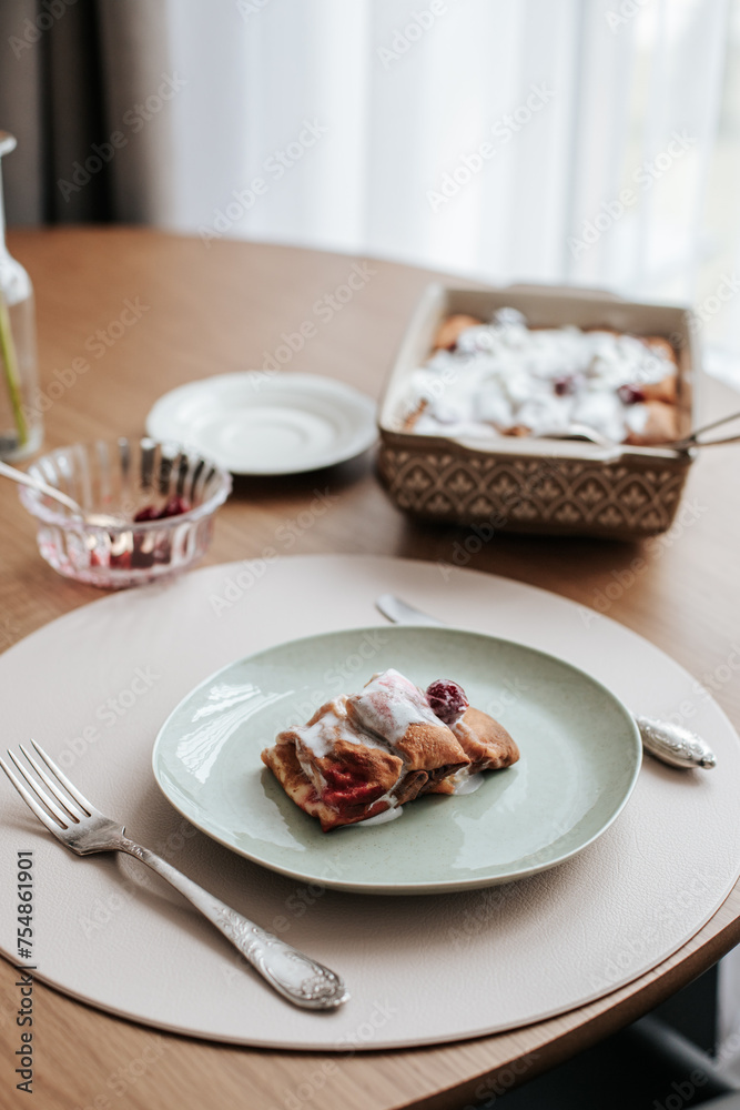Breakfast for one person is served on a round wooden table near the window. There is a cup with a saucer and tea, a dish with pancakes, ice cream and cherries. and a glass vase with a gerbera flower Breakfast for one person is served on a round wooden table near the window. There is a cup with a saucer and tea, a dish with pancakes, ice cream and cherries. and a glass vase with a gerbera flower