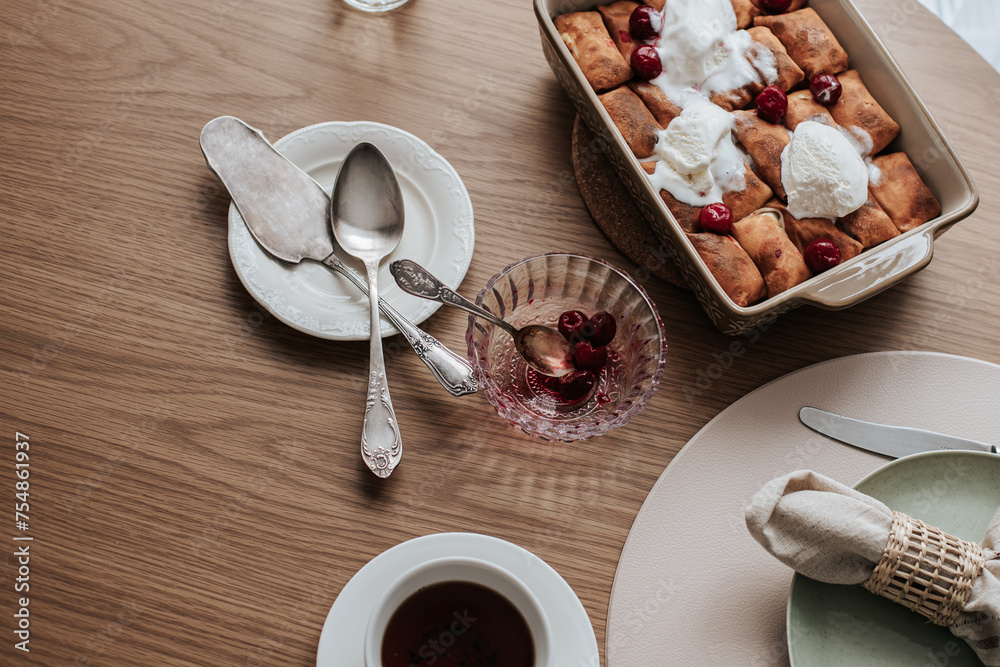 spring rolls with ice cream in a ceramic form, next to a glass bowl with cherries, a saucer with cutlery, a cup of tea and a plate with a serving napkin on a wooden table spring rolls with ice cream in a ceramic form, next to a glass bowl with cherries, a saucer with cutlery, a cup of tea and a plate with a serving napkin on a wooden table