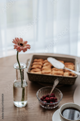 Breakfast for one person is served on a round wooden table. There is a cup with a saucer and tea  a dish with pancakes  ice cream and cherries. and a glass vase with a gerbera flower