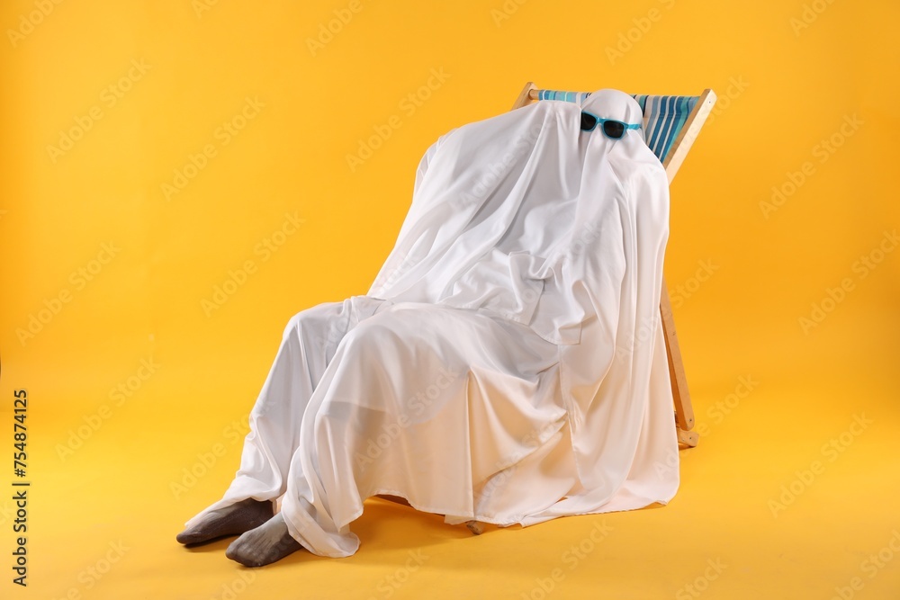 Person in ghost costume and sunglasses relaxing on deckchair against yellow background Person in ghost costume and sunglasses relaxing on deckchair against yellow background