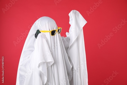 Person in ghost costume and sunglasses talking on smartphone against red background