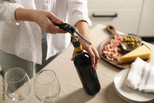 Woman opening wine bottle with corkscrew at table indoors, closeup photo
