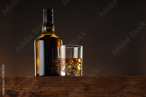 Whiskey with ice cubes in glass and bottle on wooden table, low angle view. Space for text