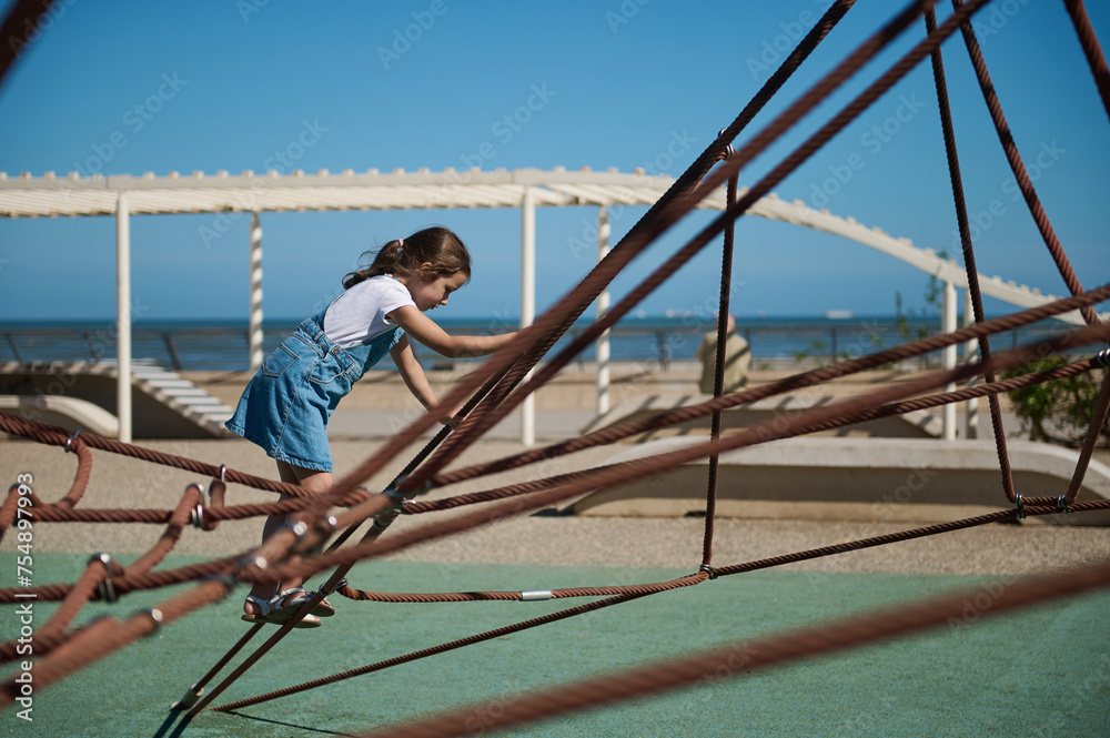 Adorable little girl climbing on a climbing net at the city playground on the embankment.