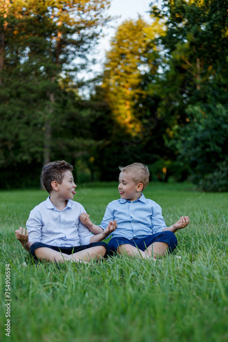 Two funny boys they sit in a yoga position, they look at each other and show their tongues, in the park.