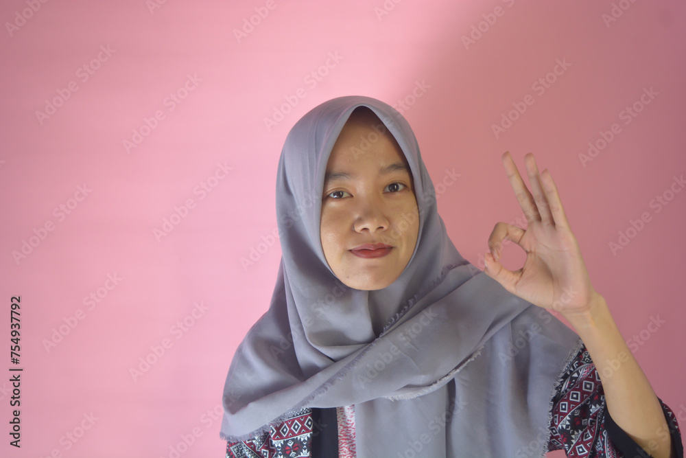 Image of young Asian woman wearing batik clothes confidently showing thumbs up in approval, recommending, liking something good, standing on pink background