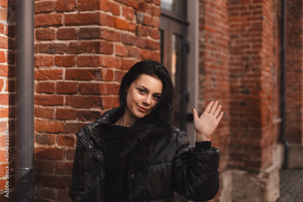 Happy attractive woman in black bomber, walk outdoors, meeting her friends or love, showing high five hey hello hi sign. She is joyful to meet her acquainted by chance. Girl wave her hand.