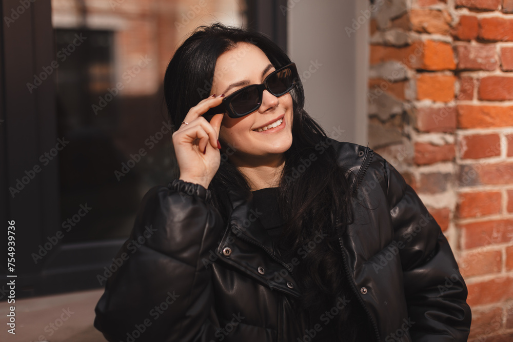 Pensive brunette woman in black puffer jacket put off black glasses and posing on office building background. Outdoor shot of happy business lady with smile look happy. Pensive brunette woman in black puffer jacket put off black glasses and posing on office building background. Outdoor shot of happy business lady with smile look happy.