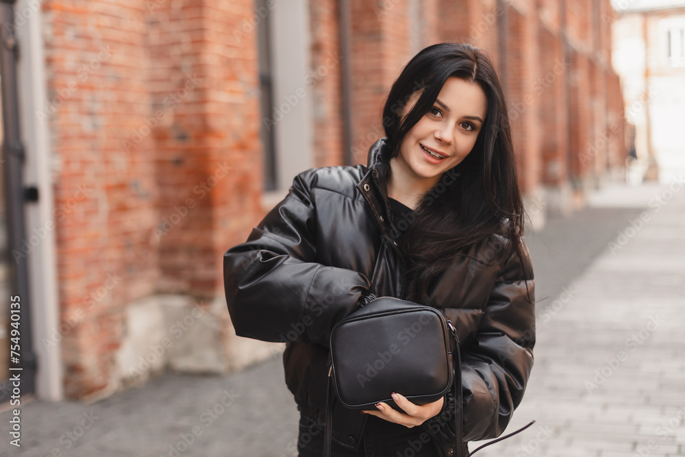 Elegant young woman looking in her black bag her phone or purse. Traveler style woman wear black puffer jacket on the street. Street style, fashion outfit.