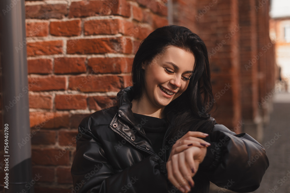 Naklejka premium Checking time. Smiling teen girl standing near brick wall background looking at her watch while waiting for her friend, read sms on her arms watch.