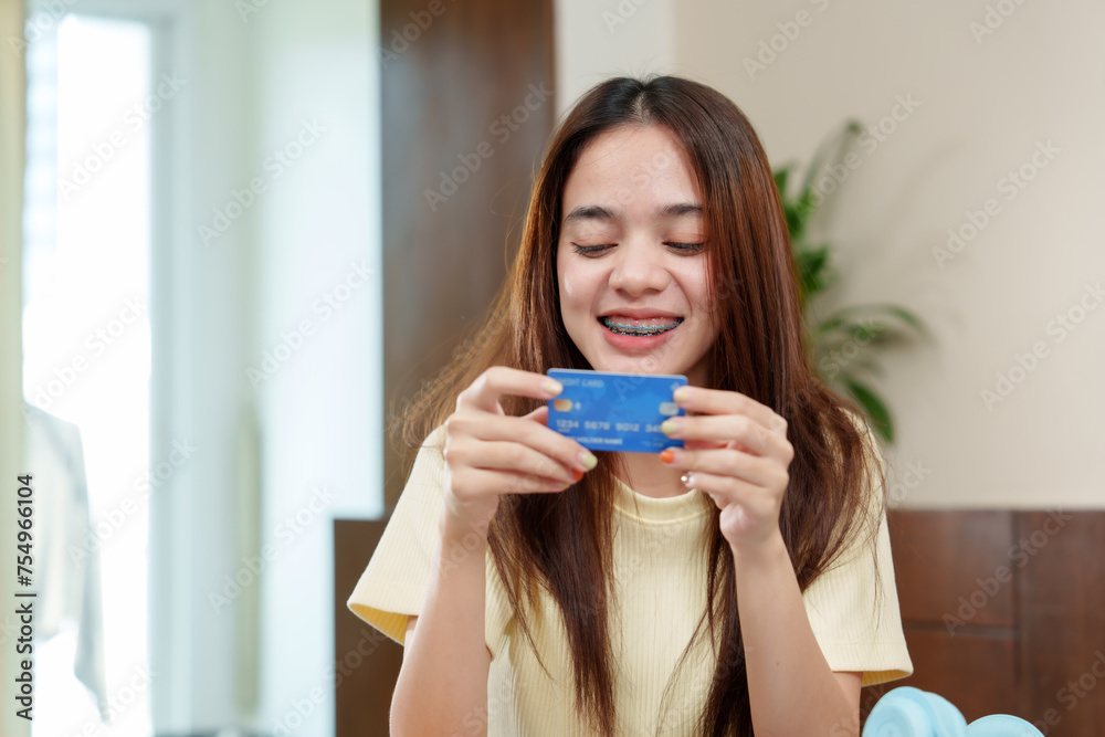 Radiant young woman with braces examines a credit card, gleeful with potential purchases, Ecstatic holds a blue credit card, anticipating an enjoyable online shopping experience at home. Radiant young woman with braces examines a credit card, gleeful with potential purchases, Ecstatic holds a blue credit card, anticipating an enjoyable online shopping experience at home.