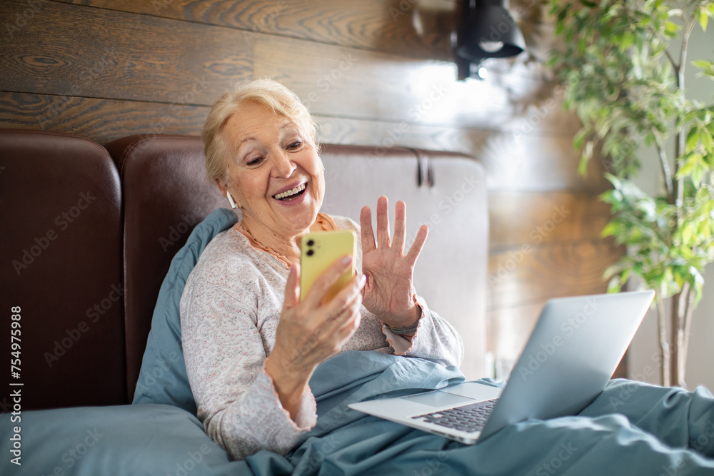 Happy elderly woman video calling on smartphone with laptop on bed Happy elderly woman video calling on smartphone with laptop on bed