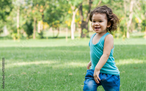 Little Caucasian little cute adorable curly hair playful girl wearing casual blue clothes, running alone with happiness and amusement in green garden or park or backyard during holidays. © Ann Rodchua