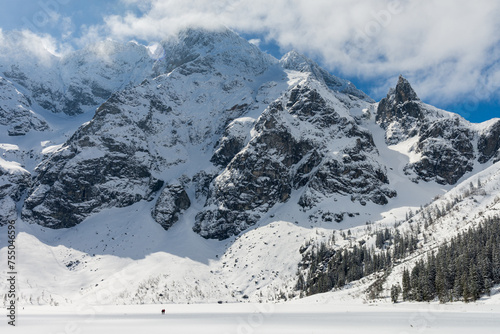 Morskie Oko Lake in Polish Tatra Mountains in sunny winter day with snowy rocky mountains in background, frozen lake and clouds