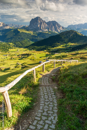 View from Seceda mountain towards the Sassolungo mouuntain in the Italian Dolomites in sunny sumer day with trekking path and wooden fence in foreground nad green grass and blue sky with clouds. photo
