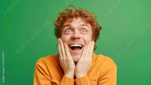 Photo of positive overjoyed young man open mouth two arms touch cheeks rejoice success isolated on green color background.
