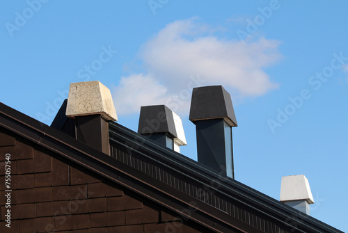 Four single and double connected dark brown to white chimneys with concrete tops covered with grey and brown metal sheet protection on top of small urban family apartment building with fluffy clouds