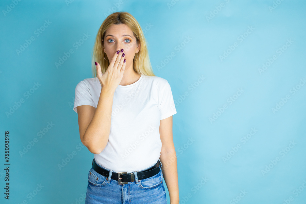 Young beautiful woman wearing casual t-shirt over isolated blue background covering mouth with hand, shocked and afraid for mistake. surprised expression