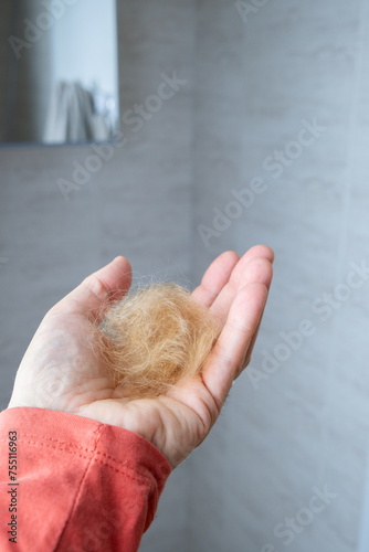The hand of a man in a red shirt holds a hairball from his pet. It is a light brown dog that has been brushed. The background is a softly out of focus bathroom.