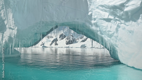 Close up iceberg arch at turquoise ocean bay with mountains on snowy shore aerial. Arctic environment preserve of ice, snow melting at global warming concept. Climate change at polar glaciers