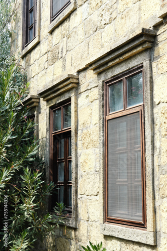 Stone Building with Windows and Lush Greenery. Tall windows with dark frames set in a textured stone wall, partially obscured by lush green oleander shrubs. Vertical photo