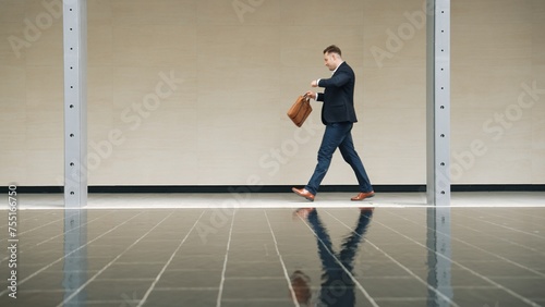 Side view of business man looking at watch while walking to train station for going to workplace. Smart caucasian manager checking time to remind for meeting and walk along modern urban city. Urbane. © Summit Art Creations