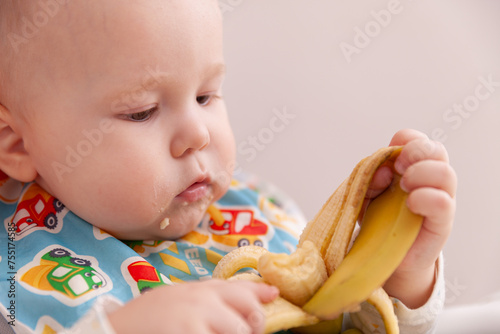 Little toddler boy in a bright bib eats banana while sitting. White gray background. Closeup