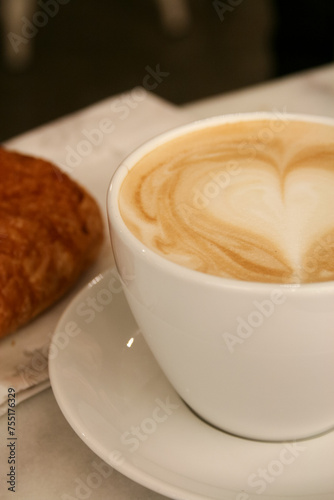 A cup of delicious morning coffee with a drawn heart and a croissant on the background. Selective soft focus