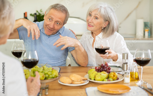 Two elderly women and elderly man chatting and drinking wine at table..