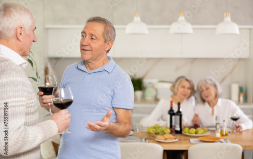 Two elderly men friends chatting and drinking wine in kitchen at home