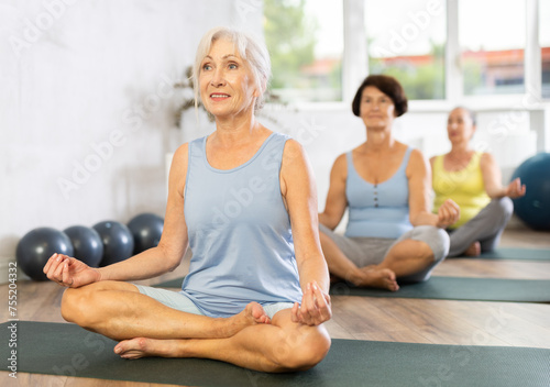 Smiling mature woman doing lotus pose of yoga on black mat in light training room