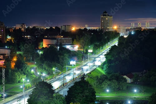Long exposure panoramic view of illuminated avenue road with moving vehicles across city green area at night