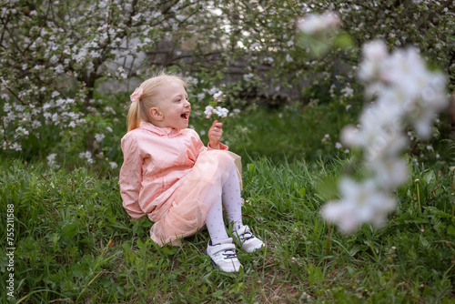 Little Caucasian girl sitting on grass in flowering garden. Child girl 4-5 years old with two ponytails wears pink dress surrounded by flowering trees.
