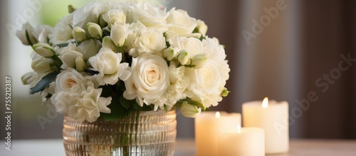 A white vase filled with fresh white flowers sits on a table next to two lit candles. The candles cast a warm glow on the elegant arrangement, creating a cozy and inviting ambiance.