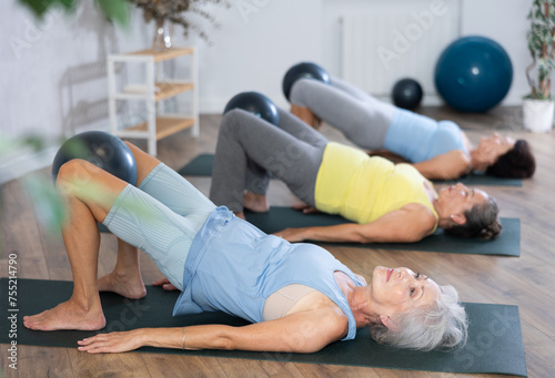 Calm old woman doing bridge pose with softball between legs together with other attendees of fitness classes