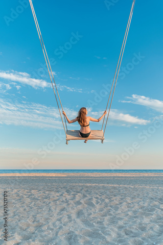 Woman in swimsuit swinging at beach over the sea on blue sky background. Vertical frame.