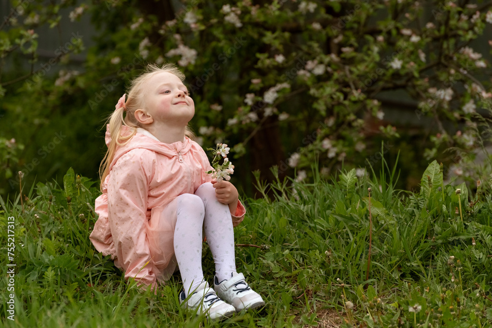 Little blond girl sitting on the ground and looking up at the sky. Child in blooming garden. Summer day Little blond girl sitting on the ground and looking up at the sky. Child in blooming garden. Summer day