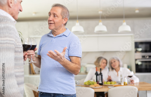 In cozy homely atmosphere, two elderly male friends have withdrawn from their female companions, stand with glass of wine in hands, communicate, discuss tips and tricks, share news