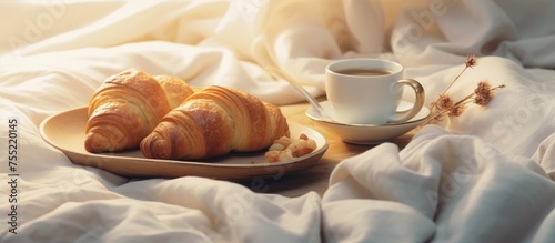 A plate with freshly baked croissants and a steaming cup of coffee sit on a white bedspread. The warm and inviting scene is perfect for a relaxing morning indulgence.