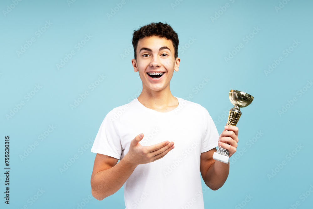 Excited boy with braces wearing white t shirt, holding trophy cup, pointing at it, looking at camera Excited boy with braces wearing white t shirt, holding trophy cup, pointing at it, looking at camera