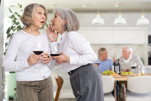 In cozy homely atmosphere, two elderly female friends have withdrawn from their male companions, stand with glass of wine in hands, communicate, discuss ladies secrets, share news photo