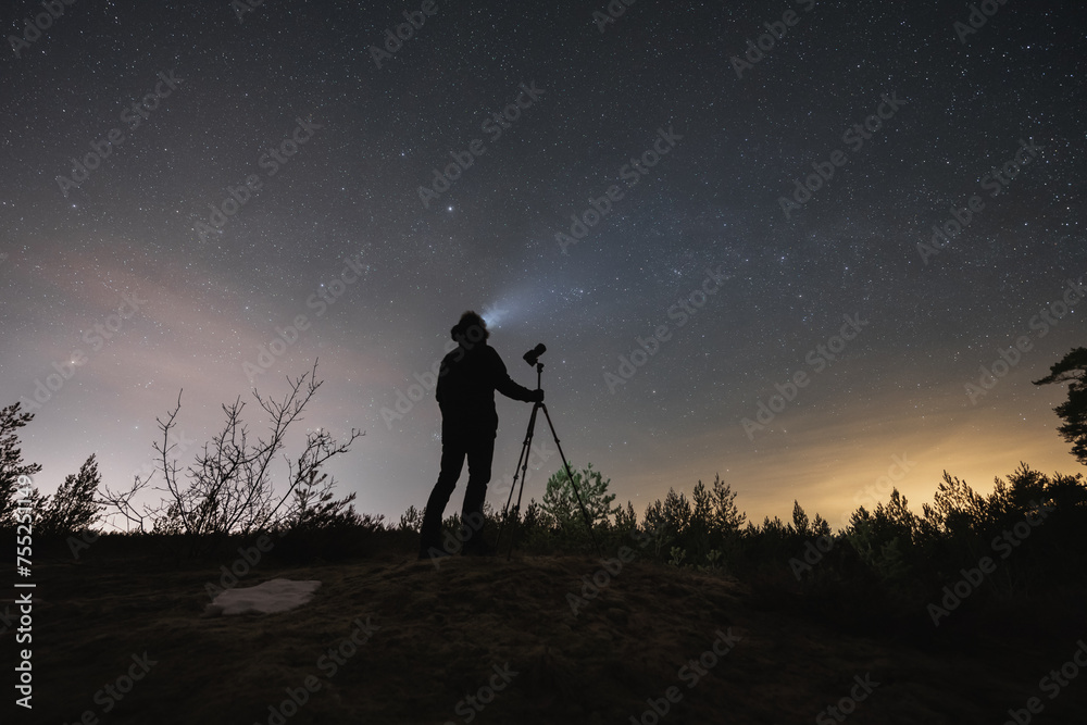 Male landscape astrophotographer with a camera on a tripod outdoors in early spring at night under the starry sky.