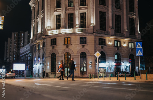 Evening stroll in the city: A couple walks past illuminated buildings on an urban street, enhancing the night's ambiance.