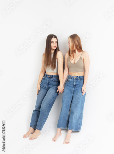 two smiling young women friends wearing jeans and tops isolated over white grey background