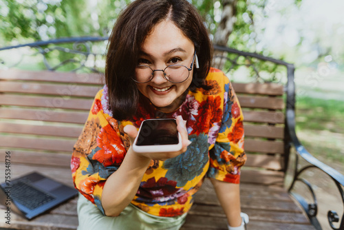 Happy woman sitting on a bench in a green park, emotionally talking on her phone. Smiling freelancer wearing a colorful blouse, animatedly chatting on her smartphone outdoor.