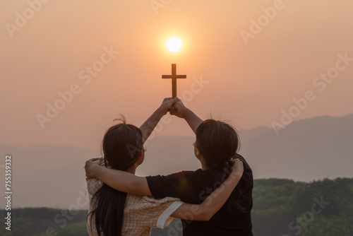 Silhouette two girl together hug with holding christian cross for worshipping God on the mountain sunset background. photo