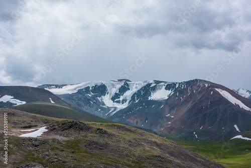 Dramatic alpine view to large snow-capped mountain range under gray rainy cloudy sky. Shadows of clouds on green stony hills and snowy rocky mountain wall. Changeable weather in high snow mountains.