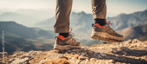 A person wearing hiking boots stands on top of a rocky mountain, surrounded by vast outdoor nature. The individual appears to be taking in the view while standing on the summit.