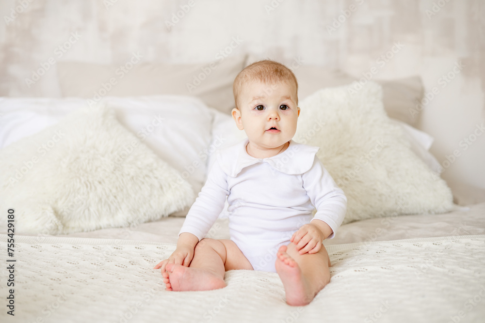 portrait of a little baby girl of six months on a bed at home in a bright bedroom smiling, happy newborn in a white cotton bodysuit